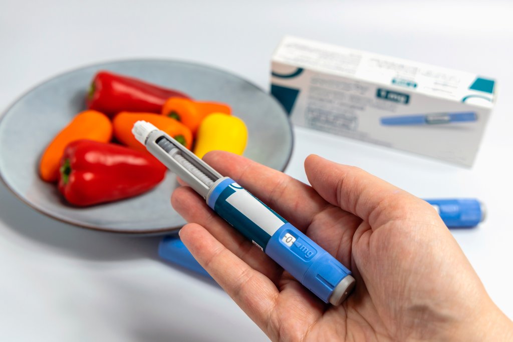 Eine Hand h&auml;lt einen blauen Abnehmspritzen-Stift. Im Hintergrund sind auf einer wei&szlig;en Fl&auml;che ein Teller mit bunten Mini-Paprikaschoten und eine Medikamentenschachtel zu sehen, die den aktuellen Abnehmen-Hype widerspiegeln.