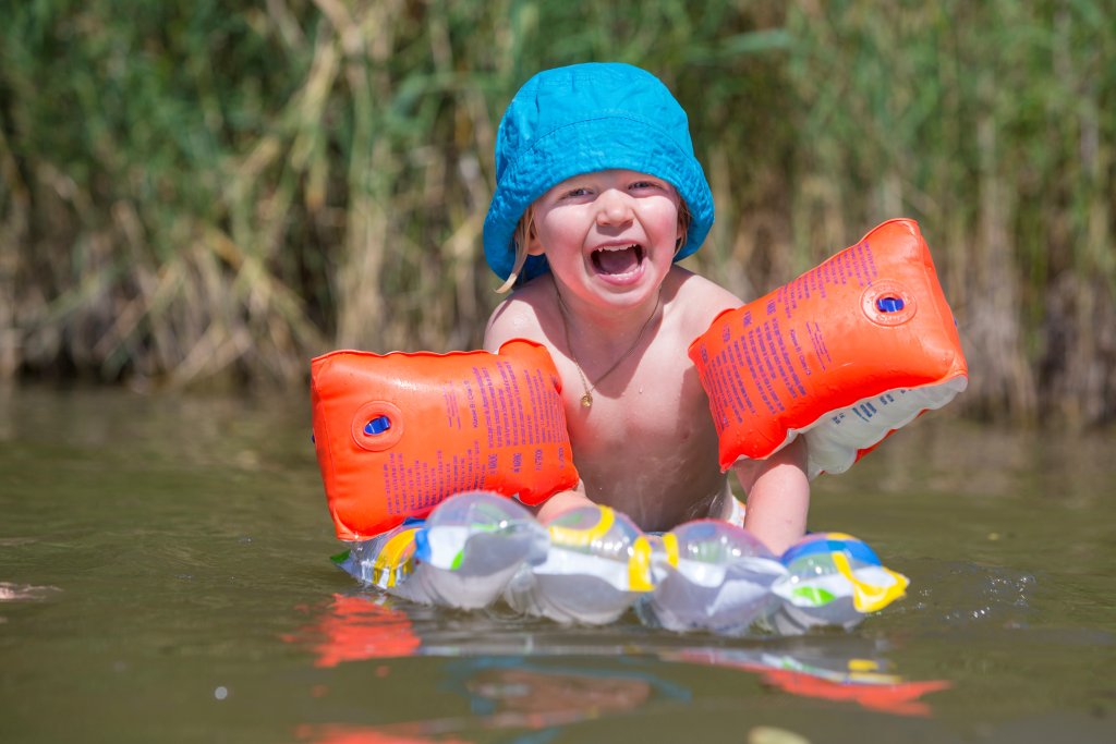 Ein l&auml;chelndes Kleinkind mit einem blauen Sonnenschutzhut und orangefarbenen Schwimmfl&uuml;geln spielt im flachen Wasser und h&auml;lt sich an einem aufblasbaren Spielzeug fest, sportlich und gesch&uuml;tzt, mit gr&uuml;nem Schilf im Hintergrund.
