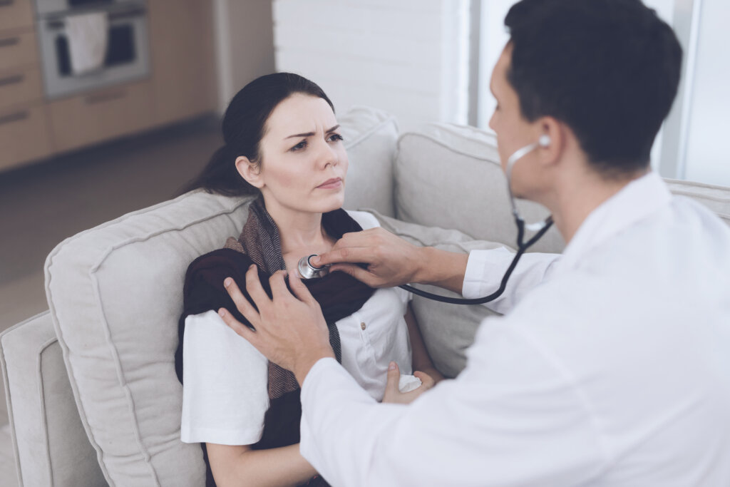 Ein Arzt h&ouml;rt mit einem Stethoskop den Brustkorb einer besorgten Frau ab, die mit einem Schal und Taschent&uuml;chern in der Hand auf einer Couch sitzt, was auf ihr Unwohlsein hindeuten k&ouml;nnte.