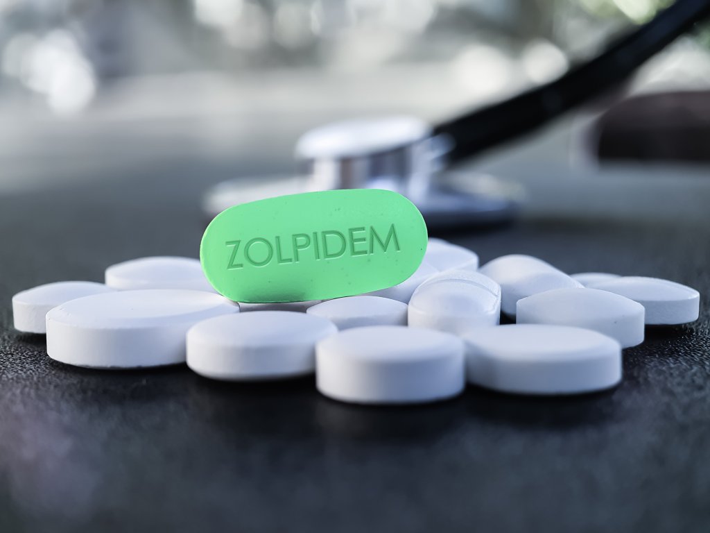 Eine gr&uuml;ne Pille mit der Aufschrift ZOLPIDEM steht aufrecht zwischen verstreuten wei&szlig;en Tabletten, mit einem Stethoskop im Hintergrund, das die Unterschiede in der Wirkung von Medikamenten f&uuml;r Frauen und kleine M&auml;nner hervorhebt.