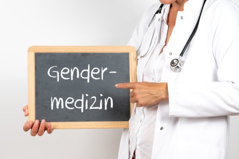 Eine Person in einem wei&szlig;en Laborkittel mit einem Stethoskop h&auml;lt eine Kreidetafel mit der Aufschrift Gender-Medizin und zeigt auf das Wort "Unterschiede zwischen M&auml;nnern und Frauen". Das Gesicht befindet sich teilweise au&szlig;erhalb des Rahmens vor einem schlichten wei&szlig;en Hintergrund.
