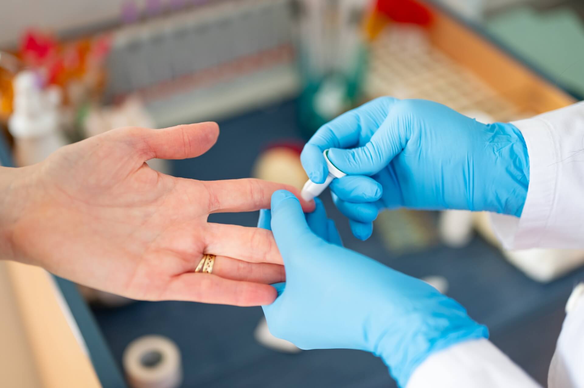 Eine medizinische Fachkraft mit blauen Handschuhen reinigt die Fingerspitze eines Patienten mit Watte, wahrscheinlich in Vorbereitung auf eine Blutuntersuchung. Der Patient tr&auml;gt einen goldenen Ring. Im Hintergrund sind medizinische Hilfsmittel zu sehen.