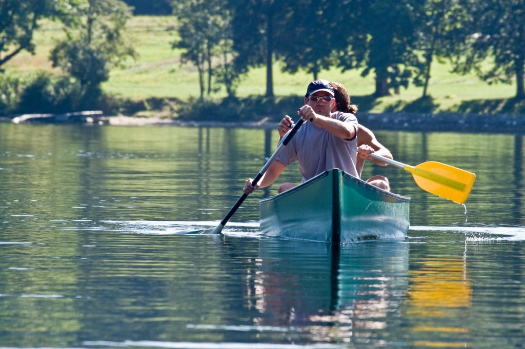 Zwei Menschen, sportlich und gesch&uuml;tzt durch Sonnenschutz, paddeln in einem gr&uuml;nen Kanu &uuml;ber einen ruhigen See, w&auml;hrend entlang des Ufers B&auml;ume und Gras zu sehen sind.