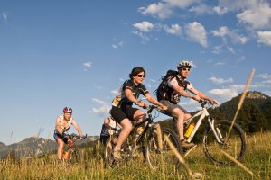Vier sportliche Radfahrer fahren mit ihren Mountainbikes auf einem grasbewachsenen Weg unter blauem Himmel und Wolkenfetzen. Berge und B&auml;ume im Hintergrund deuten auf ein gesch&uuml;tztes Outdoor-Abenteuer hin, wobei der Sonnenschutz daf&uuml;r sorgt, dass sie auf ihrer Reise bestens gesch&uuml;tzt sind.
