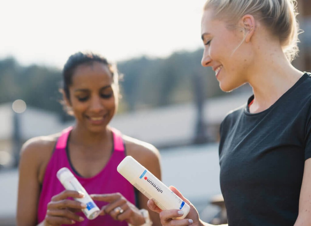 Zwei sportliche Frauen im Freien, l&auml;chelnd und mit wei&szlig;en Minisun-Flaschen in der Hand. Die eine in einem rosa Oberteil zeigt auf die Sonnenschutzflasche, w&auml;hrend die andere in einem schwarzen Oberteil zuschaut. Der Hintergrund ist unscharf.