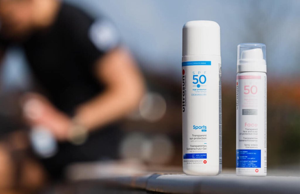 Zwei Flaschen Sonnenschutz mit der Aufschrift SPF 50 stehen auf einem Sims, im Hintergrund eine unscharfe Person, die die Lotion auftr&auml;gt - sportlich gesch&uuml;tzt, perfekt f&uuml;r Aktivit&auml;ten im Freien.