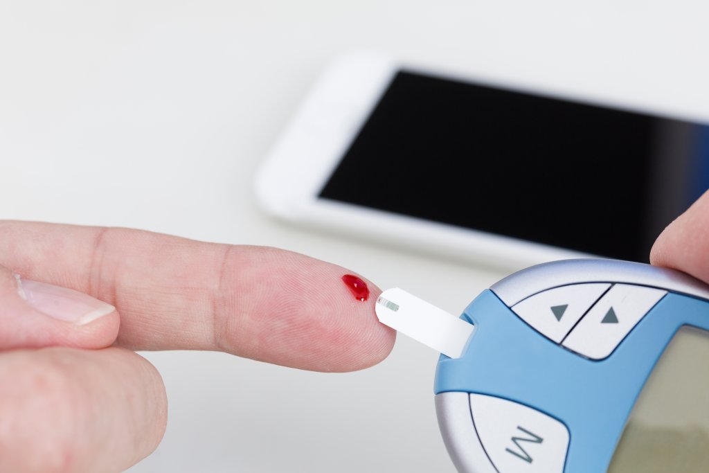 Bei einem Gesundheitscheck wird ein kleiner Blutstropfen in den Finger gestochen und auf einen Teststreifen eines Blutzuckermessger&auml;ts aufgetragen. Im Hintergrund ist ein Smartphone zu sehen - ideal f&uuml;r Selbst-Tests zuhause.