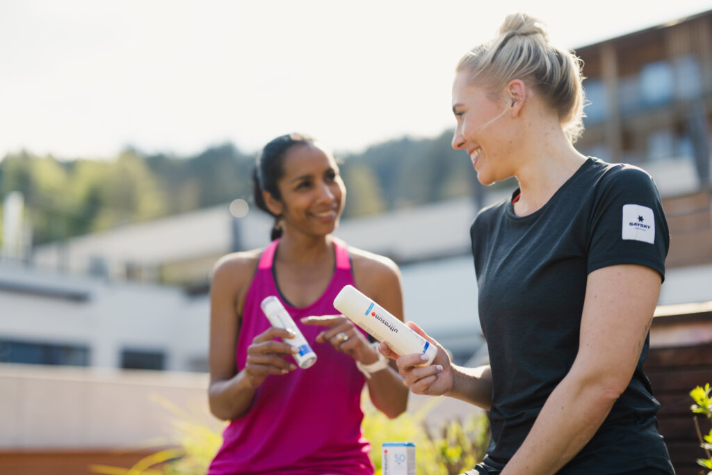 Zwei Frauen sitzen an einem sonnigen Sommertag drau&szlig;en, l&auml;cheln und halten Sonnencremeflaschen in der Hand. Die eine tr&auml;gt ein schwarzes Hemd und blondes Haar in einem Dutt, die andere ein rosa Tank-Top. Im Hintergrund sind unscharfe Geb&auml;ude und Gr&uuml;nfl&auml;chen zu sehen.