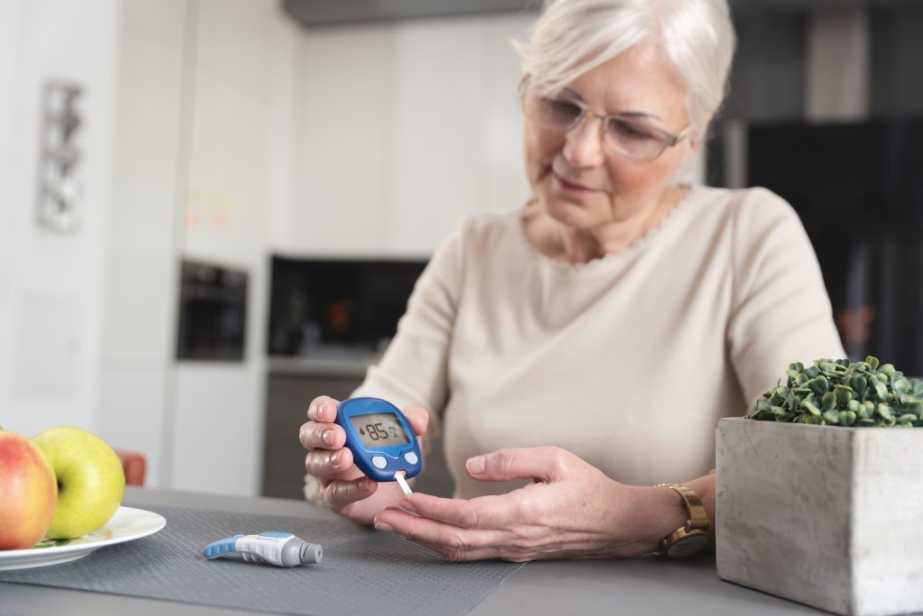 Eine &auml;ltere Frau mit grauen Haaren pr&uuml;ft ihren Blutzucker mit einem Blutzuckermessger&auml;t an einem Tisch, in dessen N&auml;he Obst und eine Topfpflanze als Teil ihrer Strategie f&uuml;r einen gesunden Lebensstil stehen.