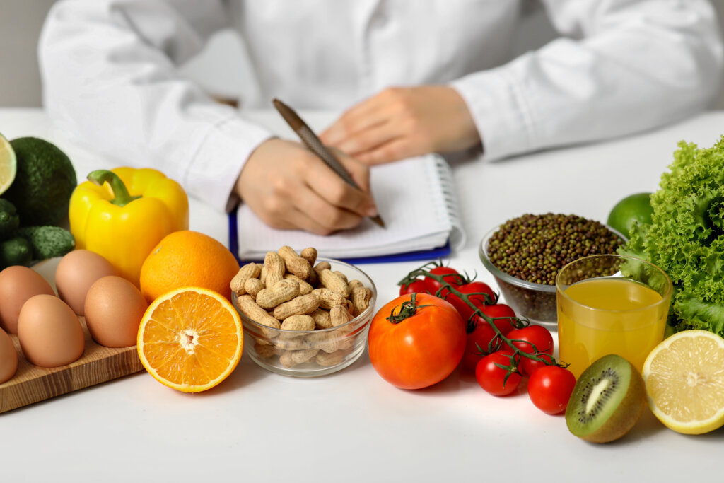Eine Person in einem wei&szlig;en Kittel schreibt in ein Notizbuch an einem Tisch mit Eiern, Erdn&uuml;ssen, Gem&uuml;se und Obst - frische Lebensmittel, die auf eine Ern&auml;hrungsplanung oder die Entwicklung einer Strategie f&uuml;r Blutzucker- und Cholesterinmanagement hindeuten.