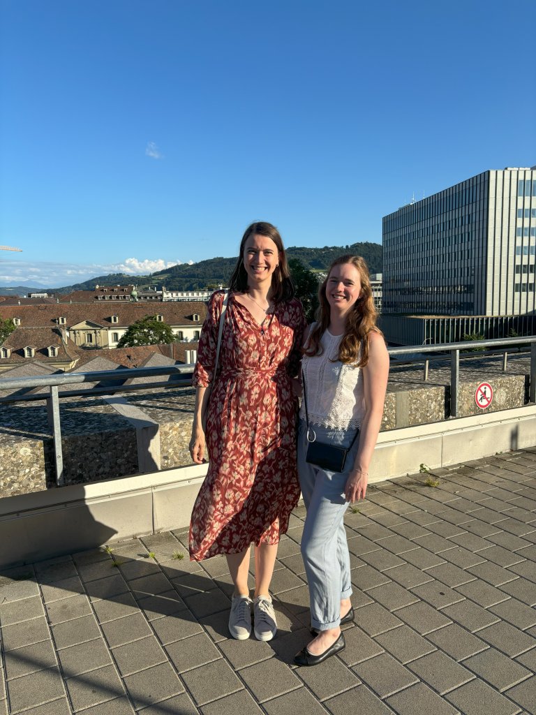 Zwei Frauen stehen l&auml;chelnd nebeneinander auf einem sonnenbeschienenen Dach - die eine, eine Fachapothekerin in einem rot gemusterten Kleid und Turnschuhen, die andere in einem wei&szlig;en Oberteil und einer hellblauen Hose. Geb&auml;ude und ein gr&uuml;ner H&uuml;gel bilden die malerische Kulisse.