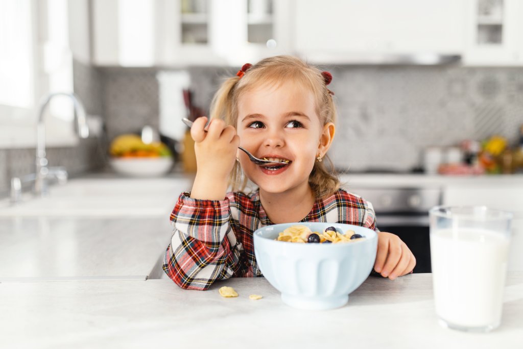 Ein junges M&auml;dchen mit Z&ouml;pfen und einem karierten Kleid l&auml;chelt, w&auml;hrend es an der K&uuml;chentheke ein angereichertes Lebensmittel-M&uuml;sli mit einem L&ouml;ffel isst. Vor ihr stehen eine Sch&uuml;ssel mit M&uuml;sli und ein Glas Milch, die beide die Gesundheit unterst&uuml;tzen.