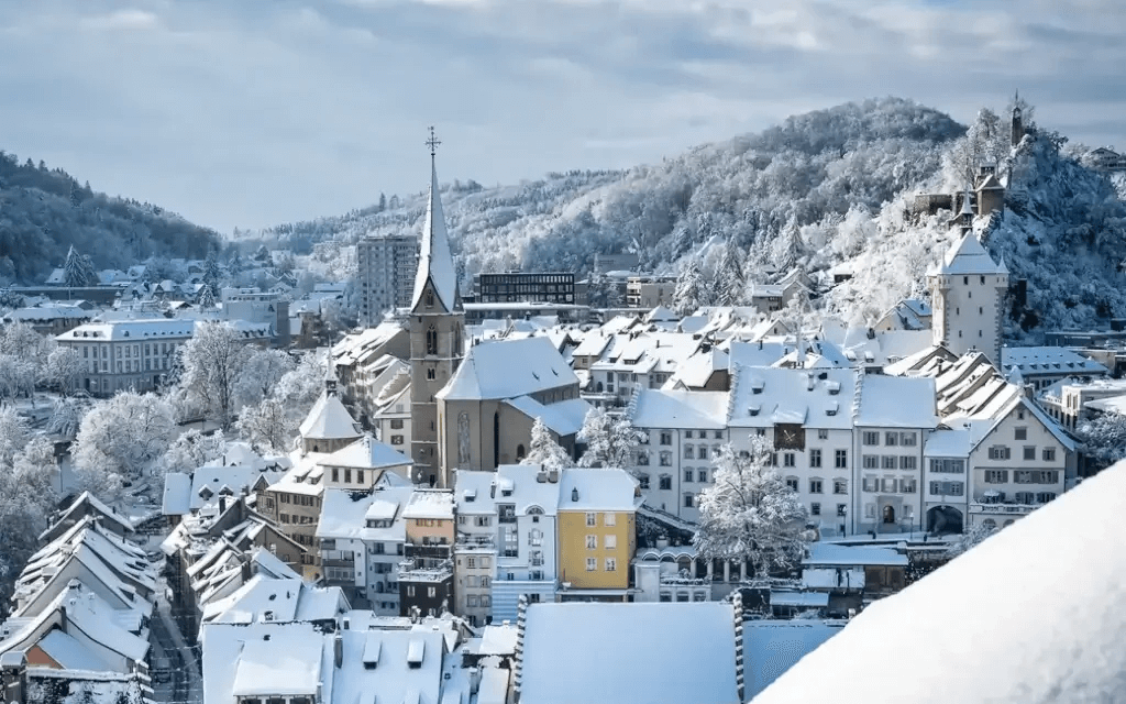 Eine malerische, verschneite europäische Stadt in der Region Baden mit einem hohen Kirchturm, bunten Gebäuden und schneebedeckten Hügeln unter einem wolkenverhangenen Winterhimmel voller Winterüberraschungen.
