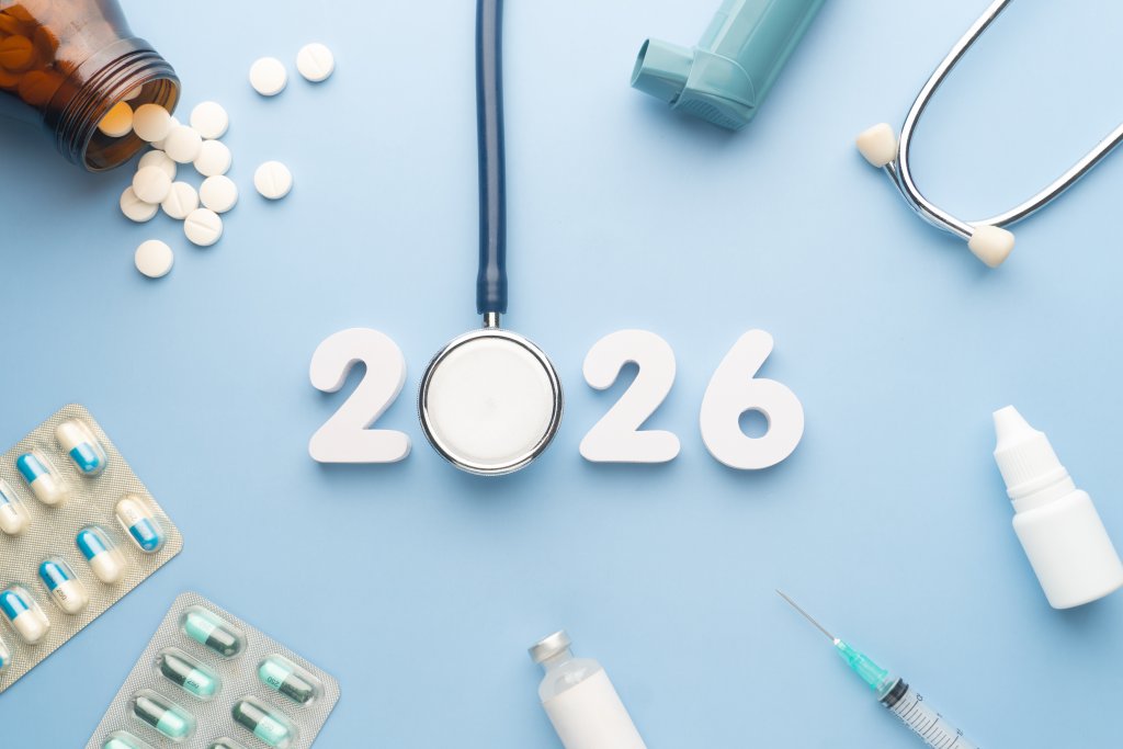 Wei&szlig;e Zahlen "2026" auf blauem Hintergrund, wobei ein Stethoskop die "0" bildet. Umliegende Gegenst&auml;nde wie Pillen, ein Pillenfl&auml;schchen, ein Inhalator und eine Spritze heben Themen wie Apotheke und LOA V 2026 im Zusammenhang mit Medikamentenpreisen hervor.