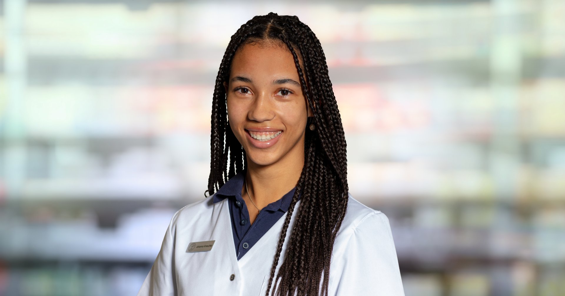 Johanna Kneschke, eine junge Frau mit langen Z&ouml;pfen, die einen wei&szlig;en Laborkittel und ein marineblaues Hemd mit Kragen tr&auml;gt, l&auml;chelt in die Kamera. Der leicht verschwommene Hintergrund deutet auf ein professionelles oder medizinisches Umfeld hin.