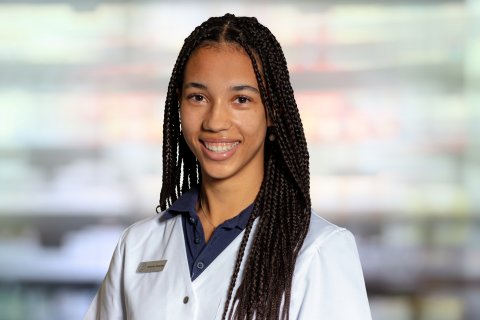 Johanna Kneschke, eine junge Frau mit langen Z&ouml;pfen, die einen wei&szlig;en Laborkittel und ein marineblaues Hemd mit Kragen tr&auml;gt, l&auml;chelt in die Kamera. Der leicht verschwommene Hintergrund deutet auf ein professionelles oder medizinisches Umfeld hin.