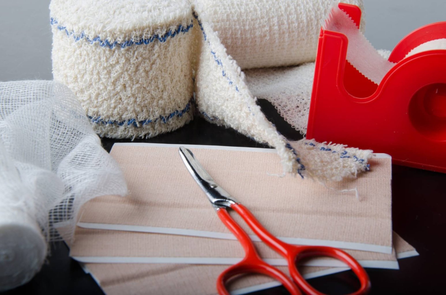 Medizinisches Zubeh&ouml;r und Sanit&auml;tsartikel wie Verbandsrollen, Pflaster, Mull, eine Schere mit rotem Griff und ein roter Klebebandabroller sind &uuml;bersichtlich auf einer schwarzen Fl&auml;che angeordnet.