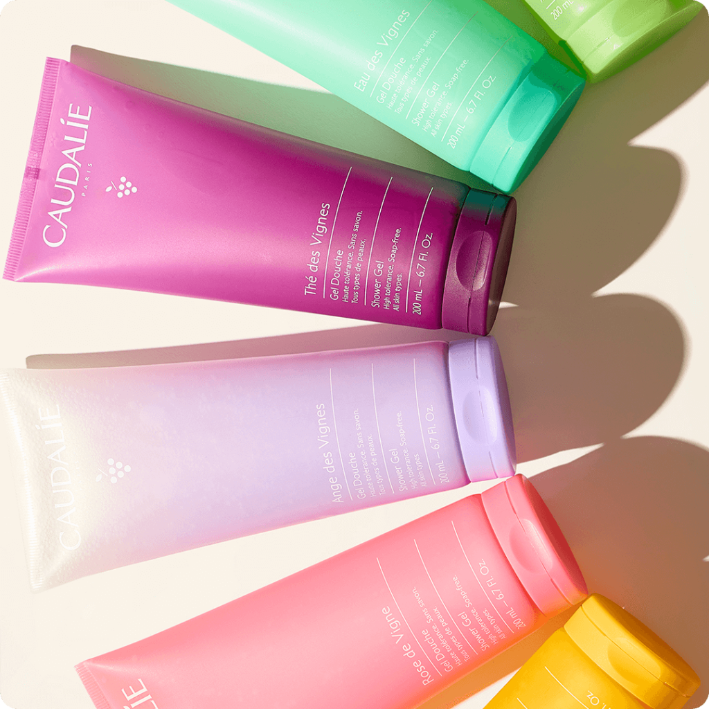 Bunte Caudalie Duschgel-Tuben (200 ml), die diagonal auf einer weißen Fläche angeordnet sind, jede in einem anderen Pastellton - rosa, lila, gelb, grün und orange - und mit ihren sanften Schatten einen lebendigen Frischekick erzeugen.