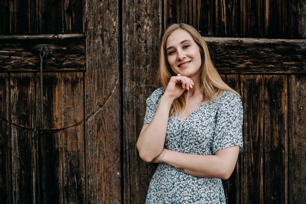 Lia Lohrer ist eine junge Frau mit blondem Haar. Sie tr&auml;gt ein hellblaues Blumenkleid und steht vor einer rustikalen Holzt&uuml;r, l&auml;chelt und st&uuml;tzt ihr Kinn auf die Hand, w&auml;hrend der Duft von Gartenblumen durch die Luft weht.