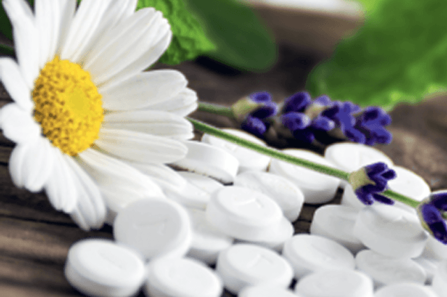 Eine Nahaufnahme von wei&szlig;en Sch&uuml;ssler-Salz-Tabletten, die auf einer h&ouml;lzernen Oberfl&auml;che verstreut sind, mit einer Kamillenbl&uuml;te, Lavendelzweigen und gr&uuml;nen Bl&auml;ttern im Hintergrund - perfekt, um diesen Fr&uuml;hling fit zu bleiben.