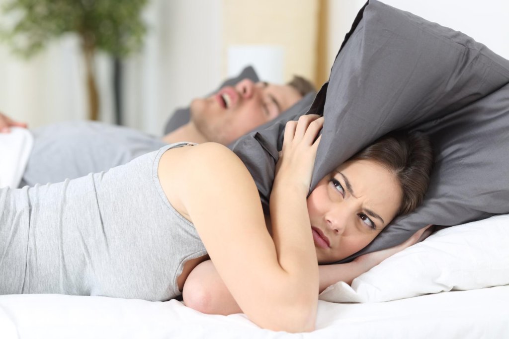 Eine Frau im Bett hält sich die Ohren mit einem Kissen zu und sieht dabei genervt aus, während ein Mann neben ihr mit offenem Mund schläft-offenbar Schnarchen auf Rezept.