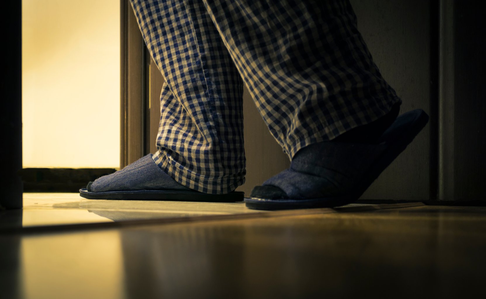Eine Person in einer blau karierten Pyjamahose und Hausschuhen betritt einen schwach beleuchteten Raum, aus dessen offener T&uuml;r ein warmes Licht str&ouml;mt &ndash; auf der Suche nach Pflanzenkraft, um bei einer Blasenentz&uuml;ndung zu helfen.